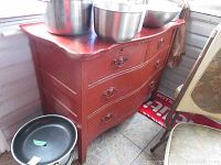 Side angle of red painted wood dresser with 4 drawers, showing surface wear, chips, and crazing. Several stainless steel bowls placed on top of dresser.