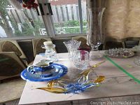 Wide view of assorted glass items on table including blue and amber decorative bowl, clear candle holder, and vintage pitcher and basin set