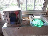 Full side view of Cuisinart rotisserie oven, Philips hand mixer in box, and Tupperware grater on table near window.