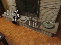 Wide photo of fireplace ledge displaying metal coffee and tea set on tray, glass serving dish, cups and saucers to left, stuffed zebra toy at far left under cups.