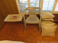 Plastic patio chair next to metal folding TV table with decorative tray top and stack of beige cushions.