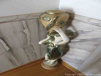 Full view of the ceramic statue showing the female figure holding a large vase with floral details against a corner wall