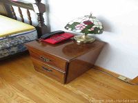 Wooden night stand with two drawers shown next to a bed, with stained glass style lamp and red telephone on top.