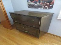 Wide view of wood dresser showing front and side with three drawers and brass and ornate metal handles; dark brown finish with some chips.