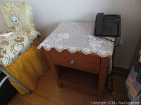 Wood night stand with lace cloth and black corded telephone on top, positioned beside a floral patterned chair.
