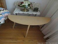 Photo of a semi-circular faux wood table with light finish next to curtain, showing three legs and wood floor beneath.