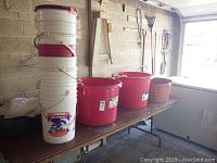 Stack of four white plastic buckets with Americana Juice Company label and red lids next to large red plastic tubs on table
