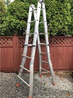 Full view of aluminum ladder standing on gravel with wooden fence background, showing construction type and adjustable height mechanism.