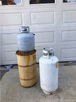 Full large white propane tank placed beside a yellow steel drum and a smaller white propane tank.