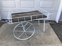 Front angled view of vintage flower cart showing white metal frame, two large spoked wheels, cedar plank bed with weathered wood, and handle on side.