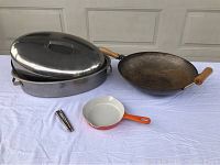 Cast iron wok, stainless steel roaster with lid, small cast iron pan, and metal nutcracker arranged on white surface