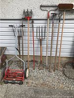 View of manual reel mower and seven wooden-handled garden tools including rakes, pitchforks, cultivators, and a broom.