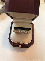 Photo of two gold-tone rings set in red leather jewelry box showing top view of rings with black enamel and rhinestones on one ring.