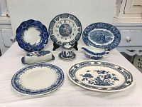 Seven items arranged on a table: large serving platter, round clock, gravy boat, lidded jar, and multiple plates all in blue and white patterns.