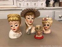 Four ceramic lady head vases displayed on a table showing different hairstyles and accessories.