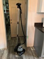 Full view of upright Conair fabric steamer with hose and steam head, placed on hardwood floor in front of door.