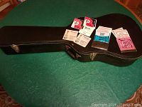 Photo of the closed black solid wooden guitar case with several guitar string packets on top, showing case size and exterior condition.