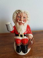 Front view showing Father Christmas holding a mug and sack, sitting on chimney base with black boots and red suit.
