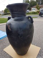 Full front view of the large black Roman style pottery vase on a cardboard base outdoors, showing the shape and handles.