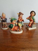 Wide shot showing all four Hummel figurines on a wood surface with neutral background.