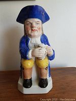Front view of a porcelain Toby jug approximately 10 inches tall, showing a seated English-style figure in a blue coat and tricorn hat holding a mug.