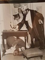 Two people in period costume at a desk, one bent under table, theatrical scene