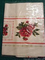 Folded beige table covering featuring red and green floral print and red and gold geometric borders, shown folded in original packaging.