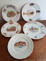 Five white antique German porcelain plates displayed on a wooden surface, each featuring nursery rhyme illustrations and German text, showing varying designs and details.