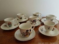 Image showing six small porcelain children's tea cups each paired with a matching saucer arranged on table, decorated with colorful illustrations of children, animals, and toys.