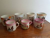 Seven antique German porcelain children's and baby mugs with cats, dogs, and sports motifs displayed on wooden surface.
