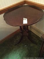 Side view of the small wooden side table in a corner showing pedestal base with three legs and scalloped edge top