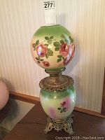 Full view of the hand painted floral glass lamp with brass details standing on a wooden surface against a wallpaper background.