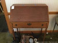 Front view of wooden secretary desk with slant-top closed and drawer closed, showing metal drawer handle and carved legs.