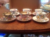 Photo of six tea cups with matching saucers arranged on table, showing variety of floral patterns and gold trim on some pieces.