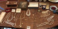 Photo showing a spread of over 50 pieces of jewelry including pearl necklaces, gold-tone necklaces, pendants, and scattered brooches and pins, all laid out on a wooden surface.