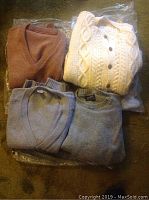 All four folded sweaters including a brown V-neck, cream cable knit cardigan, and two gray sweaters.