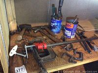 View of hand tools and clamps on wooden surface including a pipe clamp with red handle, multiple C-clamps, hammers, hand drill, pliers, and tool cans with smaller tools.
