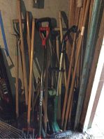 Photo showing various gardening tools including rakes, shovels, hoes, garden cultivators, and a weed trimmer clustered together in a corner.
