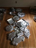 Top-down view showing aluminum pans including round, hexagonal, heart-shaped nested pans, and novelty baking pans shaped like pumpkin face, robot, star, car, Santa Claus, and an angel.