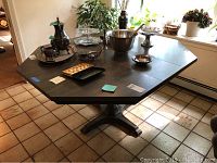 Octagonal dark wooden dining table with pedestal base and items on table surface, showing surface wear and scratches.