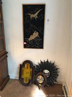 Photo shows three clocks positioned at the base of a wall near a wooden cabinet, one with yellow face and wooden frame, one with ornate sunburst plastic frame, and another wooden clock with circular face.