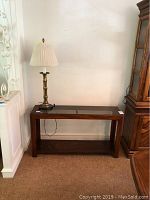 Full view of wooden console table with glass top and brass lamp on top against a white wall.