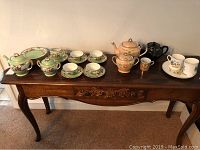 Full view of tea sets on wooden table including Noritake green set and other tea pots with mugs and plates