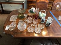 Photo showing various cut glass dishes including candy dishes, bowls, ash trays, bud vase, glass plate, and porcelain bowl along with three Norman Rockwell mugs, salt shakers, and frames arranged on a wooden table.