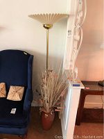 Blue wingback chair, 63 inch floor lamp with plastic pleated shade, brown vase with dried flowers and straw, seen with wall and floor.