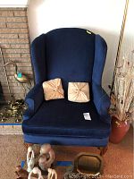Front view of navy blue wingback chair with two beige decorative pillows on seat. Carved wooden legs visible. Chair is positioned near a brick fireplace and decorative brass items.