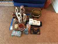 Top-down and angled view of lot displaying various figurines, trays, pottery bowls, candles, napkin holder and other decorative items arranged on carpeted floor with blue chair in background.