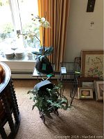 Shows black faux orchid with white flowers in a black vase on small square glass-top table; antique doll carriage with faux fern on the floor; medium glass top metal table and framed artwork in background.