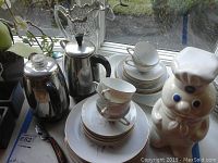 Photo showing coffee pots, stack of coffee cups and plates, and Pillsbury Doughboy cookie jar on a windowsill.