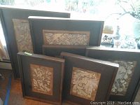 Set of six wood framed paintings laid against wall with botanical designs on cork-like paper.